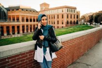 Mulher muçulmana alegre em lenço de cabeça tradicional em pé na rua da cidade e falando no telefone celular enquanto olha para longe — Fotografia de Stock