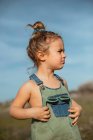 Delighted adorable little girl in overalls standing in meadow and looking away — Stock Photo