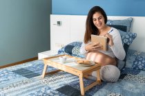 Беременная женщина сидит на кровати и пишет в блокноте во время завтрака утром — стоковое фото