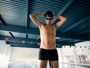 Sportsman in swimming trunks and cap with hands behind head preparing for training against pool with lanes in building — Stock Photo