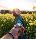 Happy young female holding boyfriend hand and looking at camera while standing on blossoming rapeseed field on sunny weather — Stock Photo