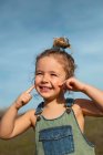 Delighted adorable little girl in overalls standing with fingers on face in meadow and looking away — Stock Photo