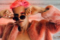 Junge Homosexuelle in stylischer Kleidung mit Tätowierung, rosa Haaren und Maniküre mit flauschiger Jacke und Sonnenbrille vor der Kamera — Stockfoto