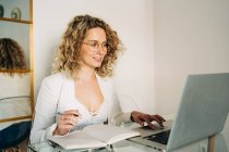 Young female freelancer with curly blond hair in casual clothes and eyeglasses taking notes in planner and looking away while working remotely using laptop at home — Foto stock