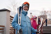 Feliz padre e hija vistiendo ropa deportiva caliente y cascos de pie con esquís en la ladera nevada de la colina y mirando la cámara con satisfacción - foto de stock