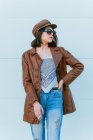 Young ethnic woman in leather jacket and sunglasses standing with hand on hip and looking away on gray background — Stock Photo