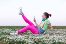 Seitenansicht Ganzkörperfreudige junge Frau in hellem Outfit liegt auf üppigem Gras mit erhobenen Beinen und surft auf dem Land mit dem Handy — Stockfoto