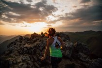 Back view of anonymous woman in activewear leaping on boulder of high ridge in valley of mountains under cloudy sky at sunset — Stock Photo