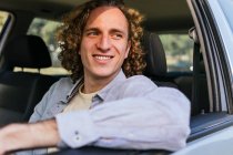 Happy young haired male looking away through open window of car while sitting at driver seat - foto de stock