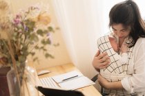 Erwachsene Mutter sitzt am Schreibtisch, arbeitet am Desktop-Computer und macht sich Notizen in Notizbuch, während sie ihr weinendes kleines Kind tagsüber am Tisch hält — Stockfoto
