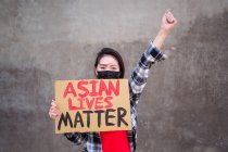 Mujer étnica en máscara y con cartel de cartón con inscripción Asian Lives Matter protestando con el brazo levantado en la calle de la ciudad y mirando a la cámara - foto de stock