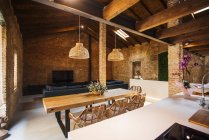 Интерьер кухни и столовой с деревянным столом и плетеными креслами под лампами против кирпичных стен в светлом доме — стоковое фото