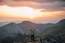 Trekker feminino com pólo e telefone em pé no pico rochoso do cume da montanha contra o céu por do sol — Fotografia de Stock