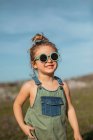 Маленькая девочка в комбинезоне и солнцезащитных очках, стоящая на лугу и наслаждающаяся летом в солнечный день в сельской местности — стоковое фото