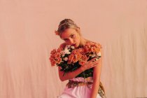 Charming romantic young bare shouldered female with bunch of fresh blooming flowers standing against beige background illuminated with neon light — Fotografia de Stock