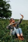 Young female in sunglasses with headphones showing peace gesture while taking self portrait on cellphone and sitting on meadow - foto de stock