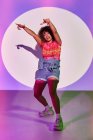 Ganzkörperfitte afroamerikanische Tänzerin in kurzen Hosen tanzt mit erhobenen Armen und ausgestreckter Zunge in die Kamera, während sie im Studio im Neonlicht steht — Stockfoto