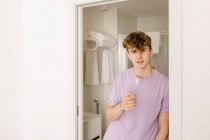 Самоуверенный молодой мужчина тысячелетия с рыжими волосами в повседневной одежде чистить зубы и смотреть в камеру, стоя в ванной комнате в солнечное утро — стоковое фото