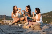 Company of young traveling female friends sitting on hill and eating at sunset in highlands — Stock Photo