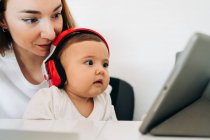 Позитивна молода мати і мила уважна дитина в навушниках дивиться мультфільм на планшеті, сидячи за столом разом — стокове фото