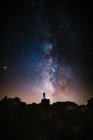 Baixo ângulo de silhueta de turista anônimo em pé com tocha de luz na cabeça em penhasco contra o céu estrelado brilhante à noite — Fotografia de Stock
