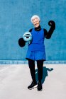Femme mûre souriante pleine longueur en vêtements de sport et gants de boxe debout avec casque à la main contre un mur bleu et regardant la caméra — Photo de stock