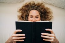 Зверху щаслива молода жінка з кучерявим волоссям лежить на ліжку читання книги під час вихідних вдома — стокове фото