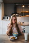 Молода жінка з ложкою і мискою насолоджується смачними кукурудзяними кільцями на кухні — стокове фото