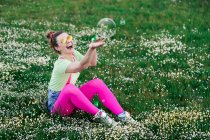 Menina jovem expressivo em roupas coloridas brilhantes brincando com bolhas de sabão e sentado no prado verde verdejante na natureza — Fotografia de Stock