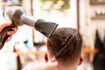 Anonymous stylist with hair dryer against man in cape in armchair in barbershop — Stock Photo