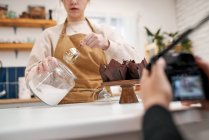 Crop fotógrafo anônimo com câmera fotográfica contra blogueiro com frasco de açúcar de confeiteiro e muffins em copos de cozimento em casa — Fotografia de Stock