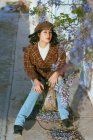 Young feminine ethnic female in stylish outfit sitting under blossoming tree branch on sunny day looking at camera — Photo de stock