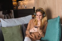 Радісна жінка сидить на дивані і насолоджується музикою в навушниках, дивлячись на екран смартфона — стокове фото