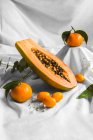 Half of pawpaw with seeds between tangerines and cumquats with leaves on white crumpled cloth — Stock Photo