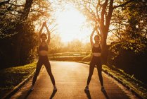 Cheerful athletic female runners standing on pathway in park and warming up arms during training at sunset — Stock Photo
