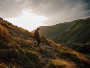 Vista laterale di femmina che cammina in discesa sul pendio roccioso della montagna in valle verde con cielo nuvoloso tramonto sullo sfondo — Foto stock