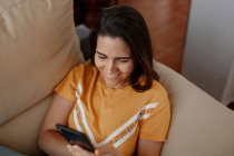 Mensajería de texto femenina joven en el teléfono celular mientras está acostado en el sofá en la sala de estar - foto de stock