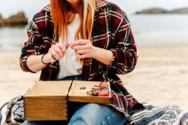 Crop anonymous craftswoman creating handmade bijouterie while using semiprecious stones and sitting on beach near sea — Stock Photo
