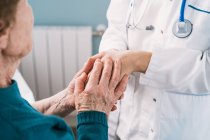 Crop médico anónimo hablando con una mujer mayor mientras se toma de la mano durante el examen en el hospital - foto de stock