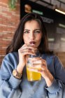Веселая молодая женщина в голубом свитере, потягивающая холодный газированный безалкогольный напиток через солому, проводя свободное время в кафетерии и счастливо глядя в камеру — стоковое фото