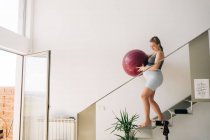 Вид збоку на молоду усміхнену майбутню жінку в спортивному одязі з м'ячем йоги, що прогулюється по сходах в будинку — стокове фото