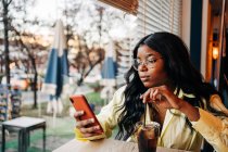 Femme afro-américaine élégante assise à table dans un café avec boisson rafraîchissante soda et la navigation sur les médias sociaux sur le téléphone mobile — Photo de stock