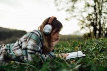 Side view of young attentive female in modern headset reading textbook while lying on meadow in summer — Fotografia de Stock