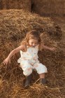 Любопытная маленькая девочка в комбинезоне спускается вниз по соломенному тюку, играя вечером в сельской местности. — стоковое фото