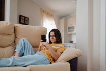 Вид сбоку молодой женщины, которая читает текст на мобильном телефоне, лежа на диване в гостиной — стоковое фото