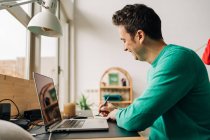 Vista lateral del alegre trabajador remoto masculino tomando notas contra netbook con pantalla negra mientras trabaja en la mesa en casa - foto de stock