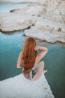 Невпізнавана жінка в стильному плаванні сидить на скелястому краю скелі і милується мальовничим морем і берегами озера, торкаючись довгого волосся. — стокове фото