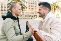 Side view of joyful young multiracial gay couple resting on wooden bench and looking at each other on city square on sunny day - foto de stock