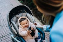 From above cute funny baby with smartphone wearing warm clothes sitting in stroller and looking at blurred mom on spring street — Stock Photo