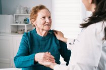 Crop médico anónimo hablando con una alegre anciana mientras se toma de la mano y se mira durante el examen en el hospital - foto de stock